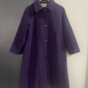 May include: A long, purple wool coat with a collared neckline and three buttons down the front. The coat has long sleeves and is hanging on a wooden hanger. The coat is a solid color and appears to be in good condition.
