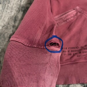 Peut inclure: Gros plan sur une manche de sweat-shirt d&eacute;lav&eacute;e de couleur rose. Le poignet c&ocirc;tel&eacute; pr&eacute;sente un aspect us&eacute; avec une petite d&eacute;chirure pr&egrave;s de la couture. Du texte d&eacute;lav&eacute; est visible sur le sweat-shirt.