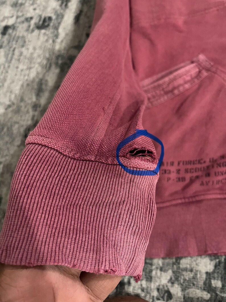 Peut inclure: Gros plan sur une manche de sweat-shirt d&eacute;lav&eacute;e de couleur rose. Le poignet c&ocirc;tel&eacute; pr&eacute;sente un aspect us&eacute; avec une petite d&eacute;chirure pr&egrave;s de la couture. Du texte d&eacute;lav&eacute; est visible sur le sweat-shirt.