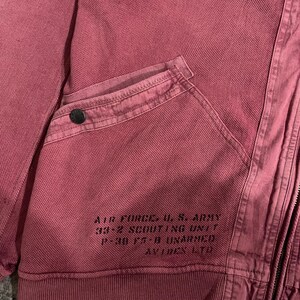 Peut inclure: Gros plan sur une veste rose poussi&eacute;reux avec un tissage textur&eacute;. Comprend une poche avec un bouton et le texte imprim&eacute; : "AIR FORCE, U.S. ARMY 33-2 SCOUTING UNIT P-38 F5-B UNARMED AVITEX LTD."