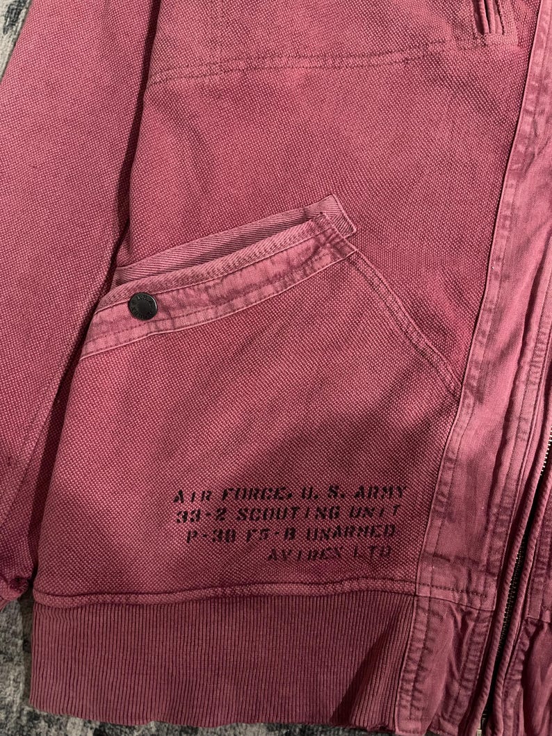 Peut inclure: Gros plan sur une veste rose poussi&eacute;reux avec un tissage textur&eacute;. Comprend une poche avec un bouton et le texte imprim&eacute; : "AIR FORCE, U.S. ARMY 33-2 SCOUTING UNIT P-38 F5-B UNARMED AVITEX LTD."