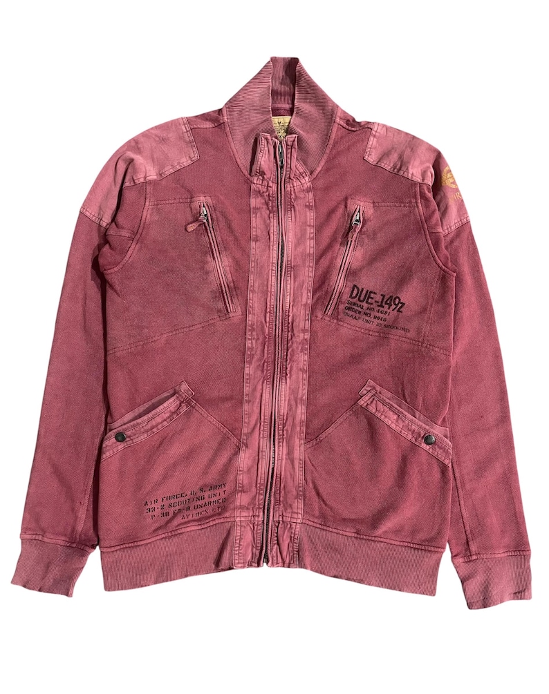 Peut inclure: Veste zipp&eacute;e bordeaux avec un col montant et plusieurs poches. La veste pr&eacute;sente du texte sur le devant et les manches, dont "DUE-149Z" et "AIR FORCE, U.S. ARMY". La veste a un aspect vintage et us&eacute;.