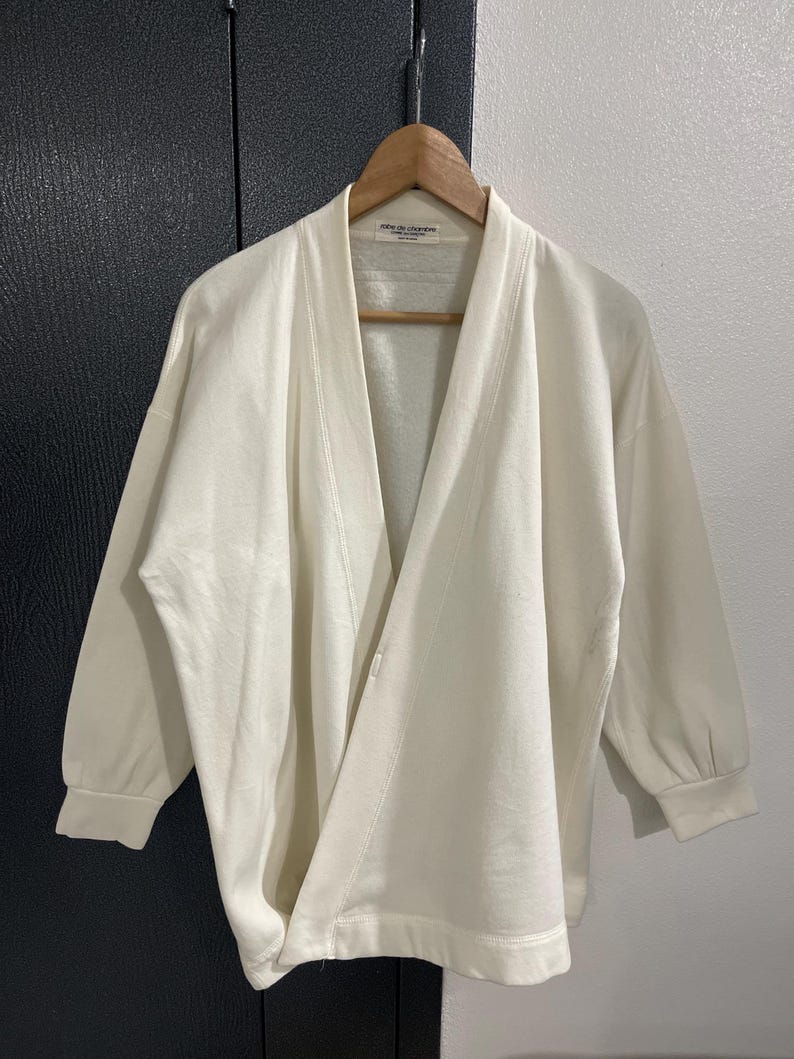 Pu&ograve; includere: Cardigan color crema con apertura frontale e maniche a tre quarti. Il cardigan &egrave; appeso a una gruccia di legno. L'indumento ha una vestibilit&agrave; morbida e un design minimalista, con dettagli di cucitura visibili.