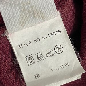 Peut inclure: Gros plan sur une &eacute;tiquette en tissu blanc avec le texte "STYLE NO.6113005" et des symboles de lavage. L'&eacute;tiquette indique que le tissu est 100% coton. Le fond est un tissu rouge fonc&eacute; textur&eacute;.
