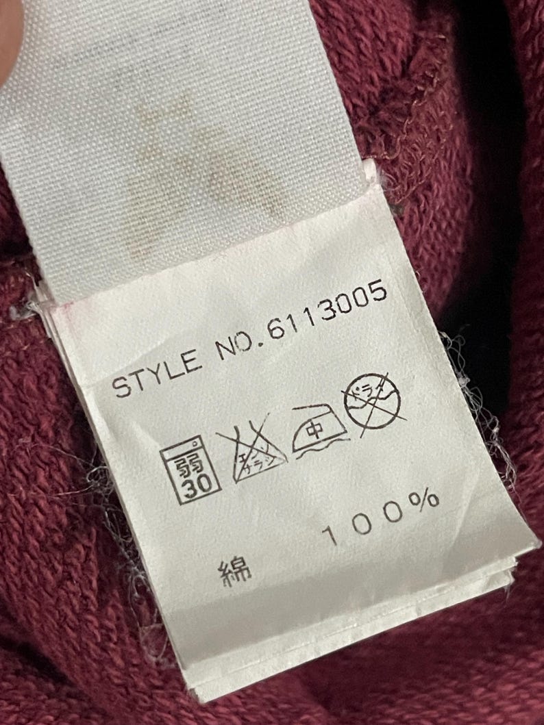 Peut inclure: Gros plan sur une &eacute;tiquette en tissu blanc avec le texte "STYLE NO.6113005" et des symboles de lavage. L'&eacute;tiquette indique que le tissu est 100% coton. Le fond est un tissu rouge fonc&eacute; textur&eacute;.