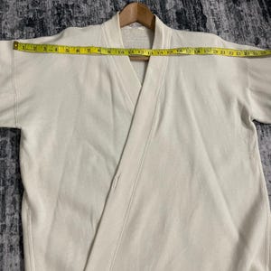 Pu&ograve; includere: Felpa color crema stile avvolgente con scollo a V e maniche corte. Il capo &egrave; misurato con un metro a nastro giallo, che mostra una larghezza di circa 63,5 cm. La felpa &egrave; appesa a una gruccia di legno.