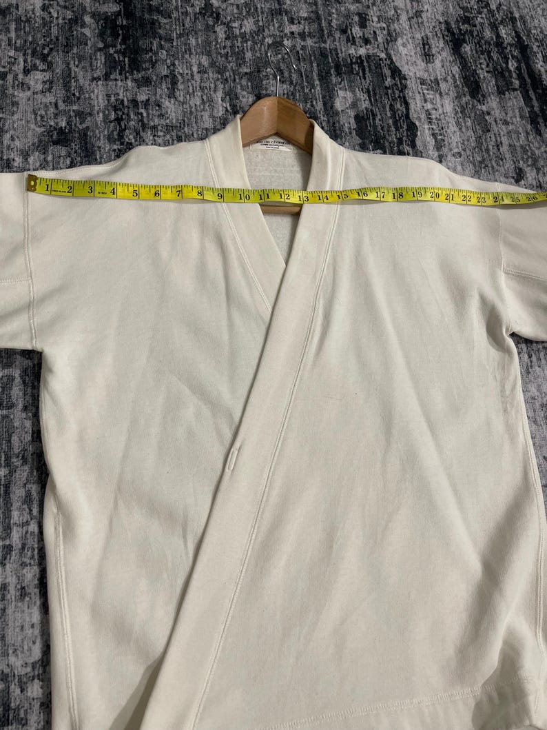 Pu&ograve; includere: Felpa color crema stile avvolgente con scollo a V e maniche corte. Il capo &egrave; misurato con un metro a nastro giallo, che mostra una larghezza di circa 63,5 cm. La felpa &egrave; appesa a una gruccia di legno.