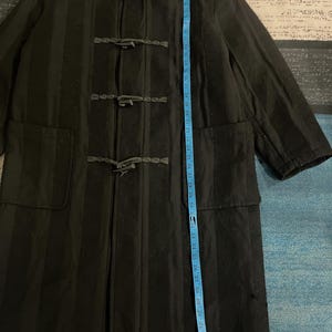 Może przedstawiać: Czarny płaszcz typu duffle z kapturem, zapięciami na kołki i dwiema przednimi kieszeniami. Płaszcz ma pionowe paski, a z boku umieszczona jest niebieska miarka. Płaszcz ma około 102 cm długości.