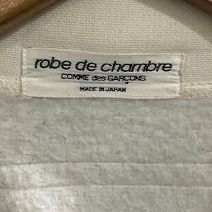 Pu&ograve; includere: Primo piano di un'etichetta di abbigliamento color crema con la scritta "robe de chambre" sopra "COMME des GARCONS" e "MADE IN JAPAN". L'etichetta &egrave; attaccata a un tessuto color crema.