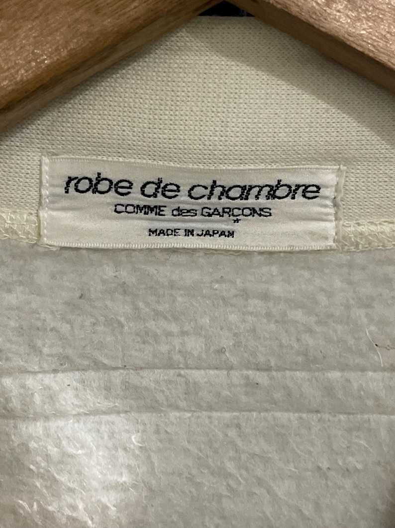 Pu&ograve; includere: Primo piano di un'etichetta di abbigliamento color crema con la scritta "robe de chambre" sopra "COMME des GARCONS" e "MADE IN JAPAN". L'etichetta &egrave; attaccata a un tessuto color crema.