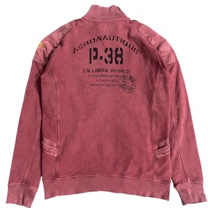 Peut inclure: Sweat-shirt zipp&eacute; rouge vieilli avec le texte "Aeronautique P.38" imprim&eacute; au dos. Le sweat-shirt pr&eacute;sente un col montant, des &eacute;cussons d'&eacute;paule et deux poches avant. Le design de style vintage comprend des d&eacute;tails d&eacute;lav&eacute;s et un aspect vieilli.