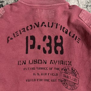 Peut inclure: Un polo rouge fan&eacute; avec un col c&ocirc;tel&eacute; et l'inscription "AERONAUTIQUE P-38 EN LISON AVIREX" en noir. Le t-shirt comporte &eacute;galement un tampon circulaire avec du texte et la date "24 OCT. 1927".