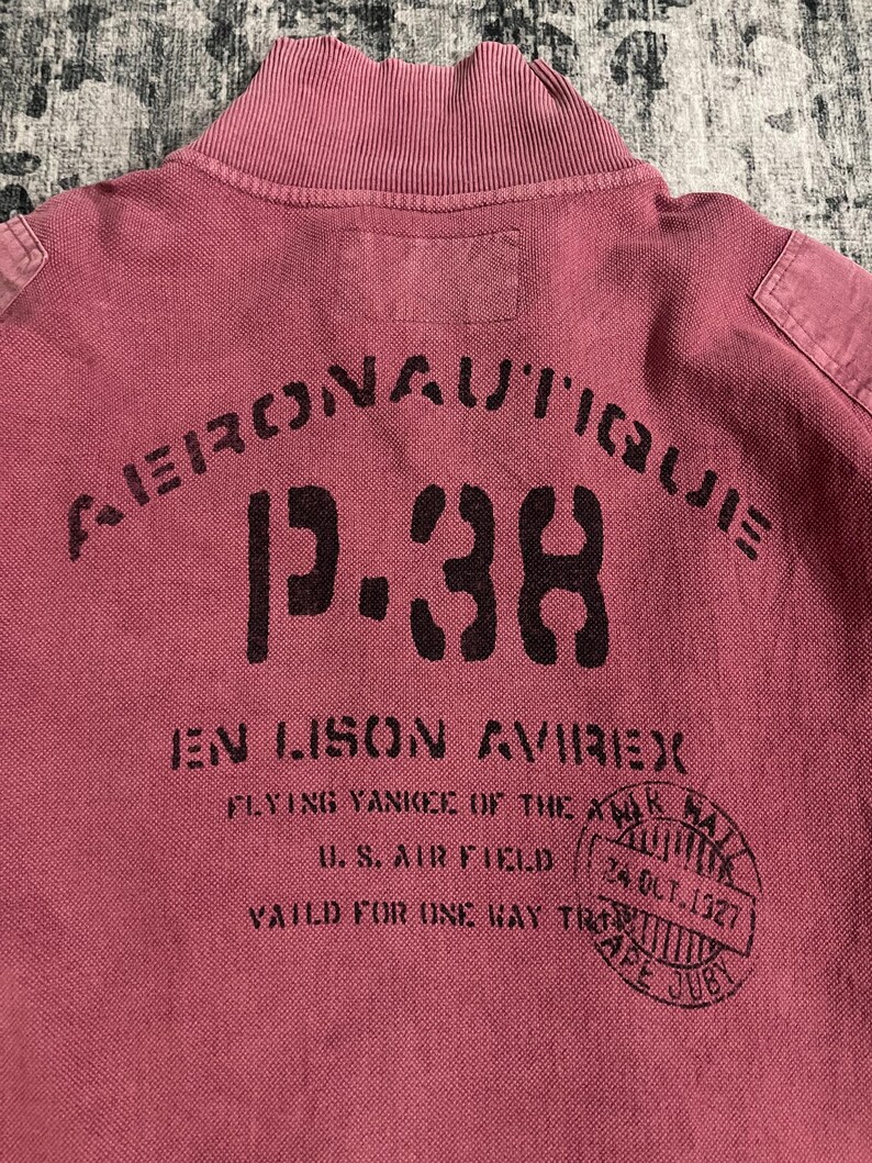 Peut inclure: Un polo rouge fan&eacute; avec un col c&ocirc;tel&eacute; et l'inscription "AERONAUTIQUE P-38 EN LISON AVIREX" en noir. Le t-shirt comporte &eacute;galement un tampon circulaire avec du texte et la date "24 OCT. 1927".
