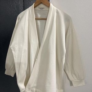 Pu&ograve; includere: Top avvolgente bianco sporco con maniche lunghe e vestibilit&agrave; morbida. Il top &egrave; appeso a una gruccia di legno. L'indumento ha uno scollo a V e una trama sottile. L'etichetta recita "robe de chambre".