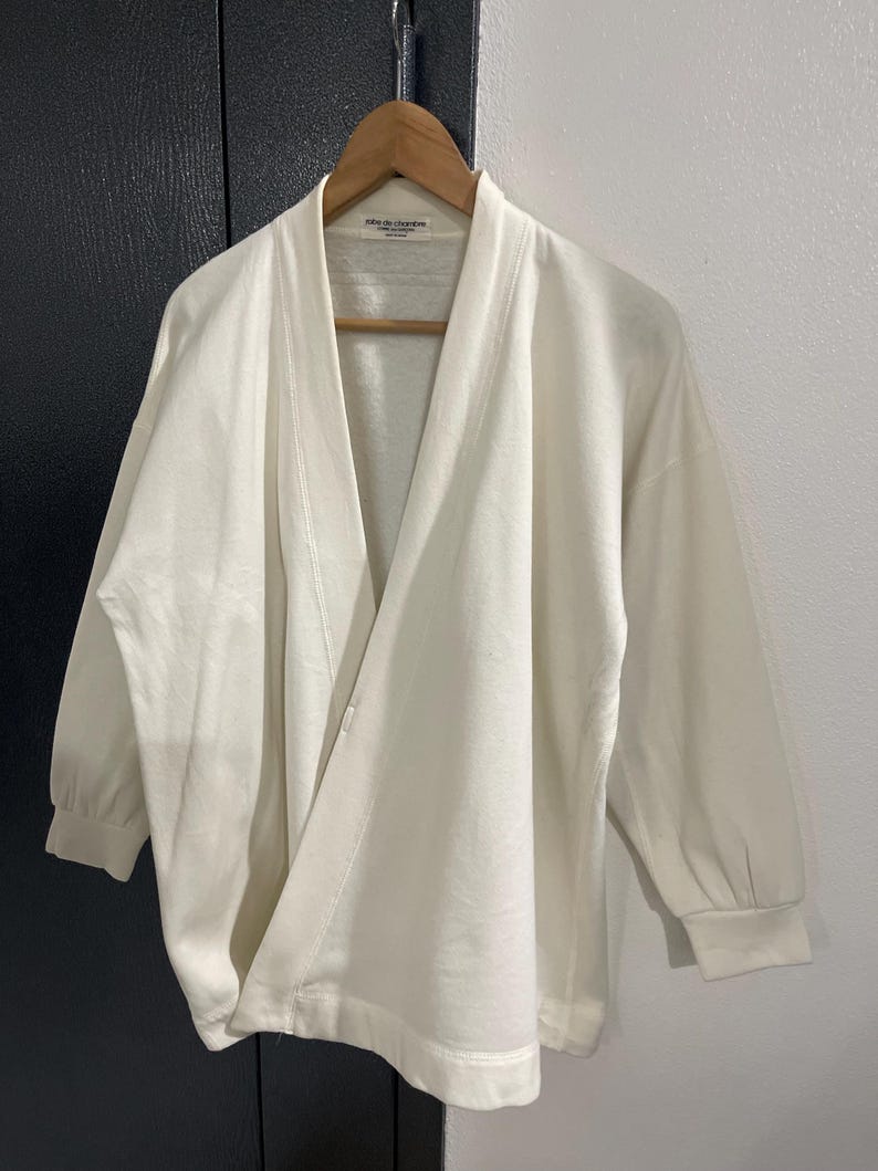 Pu&ograve; includere: Top avvolgente bianco sporco con maniche lunghe e vestibilit&agrave; morbida. Il top &egrave; appeso a una gruccia di legno. L'indumento ha uno scollo a V e una trama sottile. L'etichetta recita "robe de chambre".