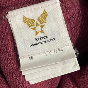 Peut inclure: Gros plan sur une &eacute;tiquette blanche avec le logo Avirex, un design &eacute;toile et ailes dor&eacute;es. L'&eacute;tiquette indique "AUTHENTIC PRODUCT" et "100%" avec du texte japonais. L'&eacute;tiquette est attach&eacute;e &agrave; un tissu bordeaux.