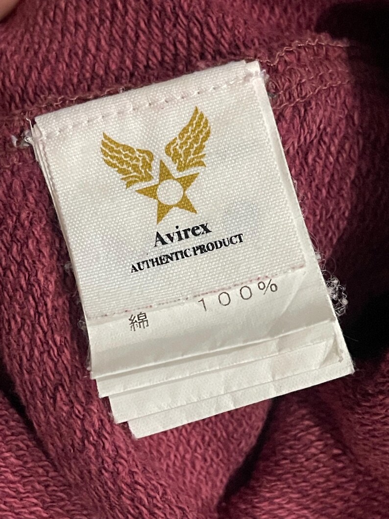 Peut inclure: Gros plan sur une &eacute;tiquette blanche avec le logo Avirex, un design &eacute;toile et ailes dor&eacute;es. L'&eacute;tiquette indique "AUTHENTIC PRODUCT" et "100%" avec du texte japonais. L'&eacute;tiquette est attach&eacute;e &agrave; un tissu bordeaux.