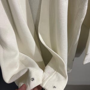 Pu&ograve; includere: Primo piano di una felpa color crema con chiusure a scatto argentate sui polsini. Il tessuto &egrave; morbido e sembra di peso medio. I polsini sono ripiegati, rivelando l'interno del capo.