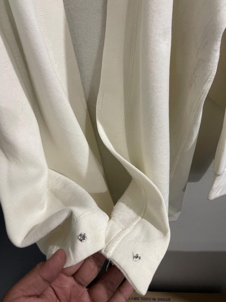 Pu&ograve; includere: Primo piano di una felpa color crema con chiusure a scatto argentate sui polsini. Il tessuto &egrave; morbido e sembra di peso medio. I polsini sono ripiegati, rivelando l'interno del capo.