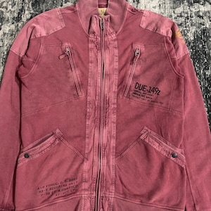 Peut inclure: Veste zipp&eacute;e rouge d&eacute;lav&eacute; avec plusieurs poches et un col montant. La veste comporte du texte sur le devant, dont "DUE-149Z" et "AIR FORCE, U.S. ARMY". La veste a un aspect vintage et us&eacute;.