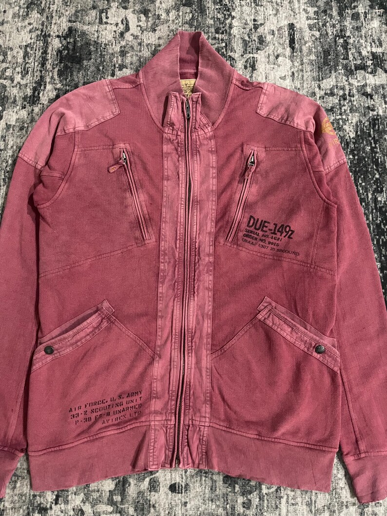 Peut inclure: Veste zipp&eacute;e rouge d&eacute;lav&eacute; avec plusieurs poches et un col montant. La veste comporte du texte sur le devant, dont "DUE-149Z" et "AIR FORCE, U.S. ARMY". La veste a un aspect vintage et us&eacute;.