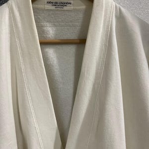 Pu&ograve; includere: Primo piano di un accappatoio color crema appeso a un'appendiabiti di legno. L'accappatoio presenta un collo a scialle e un dettaglio di cucitura sui bordi. L'etichetta recita "robe de chambre COMME des GAR&Ccedil;ONS MADE IN JAPAN."