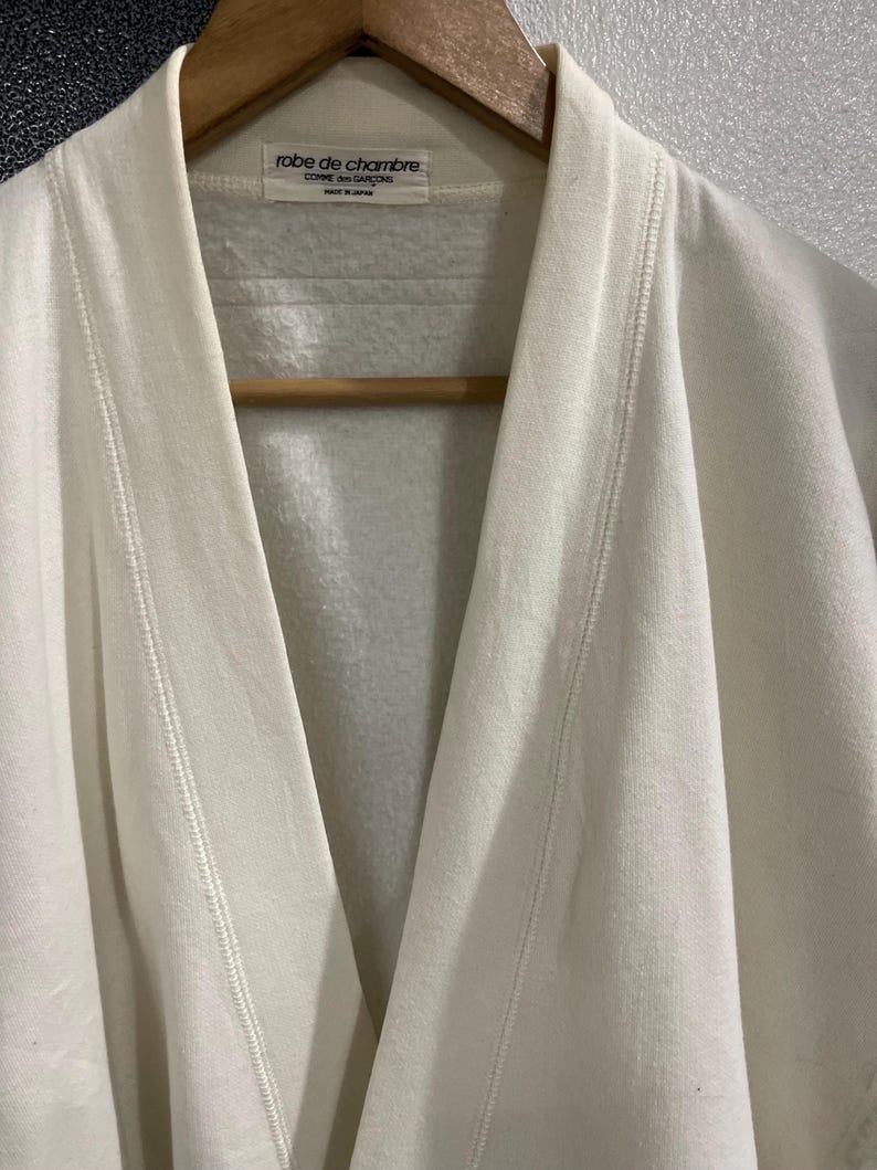 Pu&ograve; includere: Primo piano di un accappatoio color crema appeso a un'appendiabiti di legno. L'accappatoio presenta un collo a scialle e un dettaglio di cucitura sui bordi. L'etichetta recita "robe de chambre COMME des GAR&Ccedil;ONS MADE IN JAPAN."