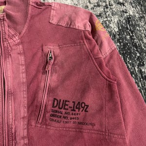 Peut inclure: Veste zipp&eacute;e rose poudr&eacute; avec col c&ocirc;tel&eacute; et empi&egrave;cements aux &eacute;paules. La veste comporte une poche poitrine avec le texte "DUE-149Z" et d'autres d&eacute;tails imprim&eacute;s. La veste a un aspect vintage et us&eacute;.