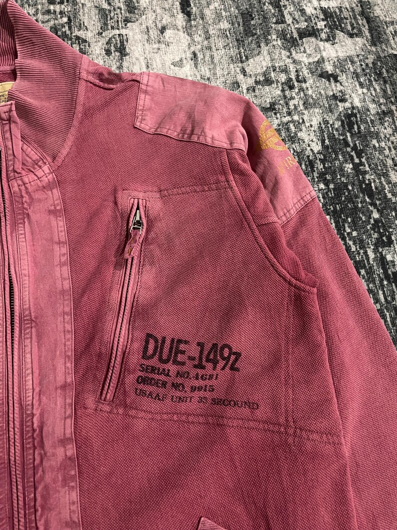 Peut inclure: Veste zipp&eacute;e rose poudr&eacute; avec col c&ocirc;tel&eacute; et empi&egrave;cements aux &eacute;paules. La veste comporte une poche poitrine avec le texte "DUE-149Z" et d'autres d&eacute;tails imprim&eacute;s. La veste a un aspect vintage et us&eacute;.