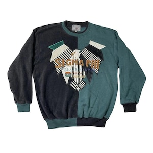 Puede incluir: Sudadera vintage con diseño dividido, mitad negra y mitad verde azulado. La parte delantera presenta un gran gráfico con el texto "SIGMA PHI" en dorado y blanco, con un diseño similar a un águila. Los puños y el cuello son acanalados.