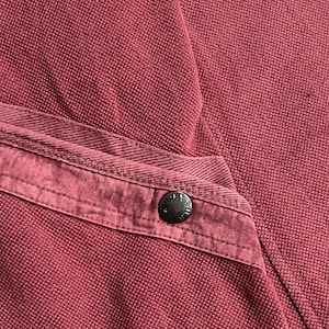 Peut inclure: Gros plan sur un v&ecirc;tement bordeaux, possiblement une chemise ou une veste, avec un tissu textur&eacute;. Un bouton fonc&eacute; est visible sur un d&eacute;tail de poche. Le bouton a un design circulaire.