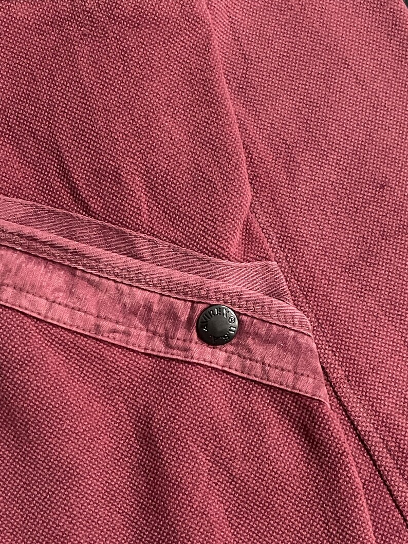 Peut inclure: Gros plan sur un v&ecirc;tement bordeaux, possiblement une chemise ou une veste, avec un tissu textur&eacute;. Un bouton fonc&eacute; est visible sur un d&eacute;tail de poche. Le bouton a un design circulaire.