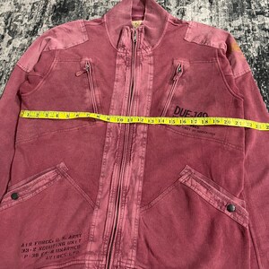 Peut inclure: Veste zipp&eacute;e rouge d&eacute;lav&eacute; avec plusieurs poches et un col c&ocirc;tel&eacute;. La veste comporte le texte "AIR FORCE U.S. ARMY 33-2 SCOUTING UNIT P-30 F-4 UNARMED AVIREX LTD" et "DUE-140". La veste mesure environ 58 cm de long.