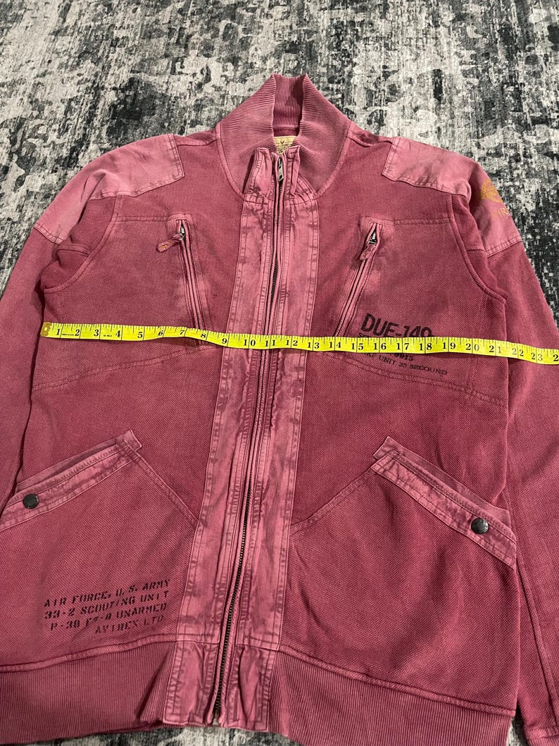Peut inclure: Veste zipp&eacute;e rouge d&eacute;lav&eacute; avec plusieurs poches et un col c&ocirc;tel&eacute;. La veste comporte le texte "AIR FORCE U.S. ARMY 33-2 SCOUTING UNIT P-30 F-4 UNARMED AVIREX LTD" et "DUE-140". La veste mesure environ 58 cm de long.