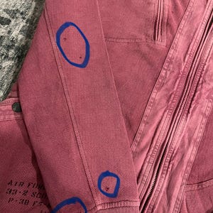 Peut inclure: Gros plan sur une veste zipp&eacute;e rose poussi&eacute;reux. La veste pr&eacute;sente un poignet c&ocirc;tel&eacute; et une fermeture &eacute;clair. La manche gauche porte l'inscription "AIR FORCE 33-2 SQD P-30 F". Le tissu a un aspect textur&eacute;.