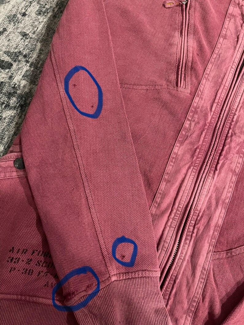 Peut inclure: Gros plan sur une veste zipp&eacute;e rose poussi&eacute;reux. La veste pr&eacute;sente un poignet c&ocirc;tel&eacute; et une fermeture &eacute;clair. La manche gauche porte l'inscription "AIR FORCE 33-2 SQD P-30 F". Le tissu a un aspect textur&eacute;.