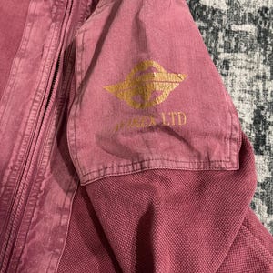 Peut inclure: Veste rose poussi&eacute;reux avec fermeture &eacute;clair. Le haut de la manche pr&eacute;sente un logo dor&eacute; et le texte "VIXON LTD". Le tissu a un aspect textur&eacute;. La veste est faite d'un mat&eacute;riau robuste.