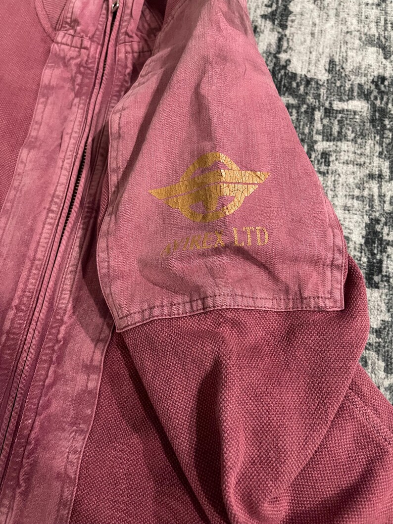 Peut inclure: Veste rose poussi&eacute;reux avec fermeture &eacute;clair. Le haut de la manche pr&eacute;sente un logo dor&eacute; et le texte "VIXON LTD". Le tissu a un aspect textur&eacute;. La veste est faite d'un mat&eacute;riau robuste.