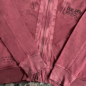 Peut inclure: Veste &agrave; capuche zipp&eacute;e rouge d&eacute;lav&eacute; avec plusieurs poches et poignets c&ocirc;tel&eacute;s. La veste est dot&eacute;e d'une fermeture &eacute;clair int&eacute;grale et d'un texte sur la manche sup&eacute;rieure droite qui se lit "DUE-149Z". La veste porte &eacute;galement le texte "AIR FOR" sur la manche gauche.