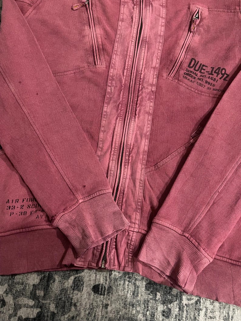 Peut inclure: Veste &agrave; capuche zipp&eacute;e rouge d&eacute;lav&eacute; avec plusieurs poches et poignets c&ocirc;tel&eacute;s. La veste est dot&eacute;e d'une fermeture &eacute;clair int&eacute;grale et d'un texte sur la manche sup&eacute;rieure droite qui se lit "DUE-149Z". La veste porte &eacute;galement le texte "AIR FOR" sur la manche gauche.