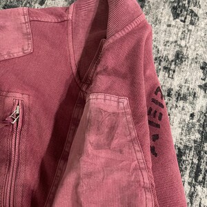 Peut inclure: Veste zipp&eacute;e bordeaux avec col et poignets c&ocirc;tel&eacute;s. La veste pr&eacute;sente un tissu textur&eacute; et une poche plaqu&eacute;e sur le haut du bras. La manche porte des lettres noires. C'est un v&ecirc;tement d'ext&eacute;rieur d&eacute;contract&eacute;.