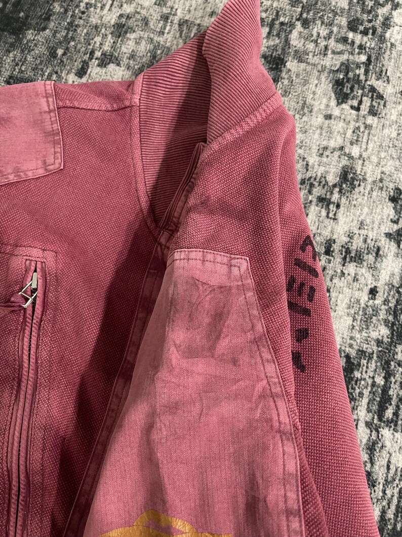 Peut inclure: Veste zipp&eacute;e bordeaux avec col et poignets c&ocirc;tel&eacute;s. La veste pr&eacute;sente un tissu textur&eacute; et une poche plaqu&eacute;e sur le haut du bras. La manche porte des lettres noires. C'est un v&ecirc;tement d'ext&eacute;rieur d&eacute;contract&eacute;.