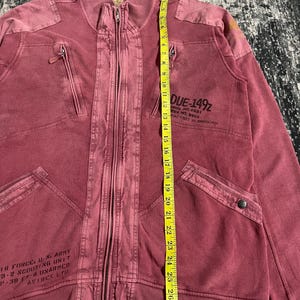 Peut inclure: Veste zipp&eacute;e bordeaux avec plusieurs poches et un col c&ocirc;tel&eacute;. La veste comporte du texte &agrave; l'avant et un m&egrave;tre ruban jaune. La veste mesure environ 74 cm de long.