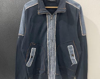Op de afbeelding: Een marineblauwe jas met ritssluiting en denim accenten op de schouders, zakken en in het midden. De jas heeft een geribbelde zoom en manchetten, en een klein logo-label. De jas hangt aan een houten hanger.