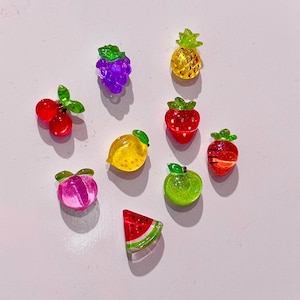 Puede incluir: Surtido de charms de resina con forma de fruta de colores, que incluyen una piña, uvas, cerezas, fresas, un limón, una manzana verde, un melocotón y una rodaja de sandía.