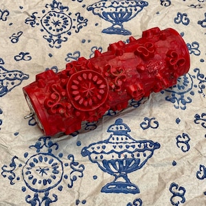 Könnte beinhalten: Eine leuchtend rote, dekorative Walze mit aufwendigen floralen und kreisförmigen Designs. Die Walze besteht aus einem festen Material und hat einen Metallstift. Sie liegt auf einem gemusterten Papier mit blau-weißen Designs.