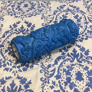 Pode incluir: Um rolo de pintura azul com um padrão floral detalhado. O rolo é feito de um material duro e tem um eixo de metal. Está sobre uma superfície branca com um desenho floral azul.