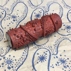 Könnte beinhalten: Eine rote Farbwalze mit Muster und Metallgriff liegt auf gemustertem Papier. Die Walze hat eine strukturierte Oberfläche mit verschiedenen Formen und Designs. Das Papier hat einen weißen Hintergrund mit blauen Paisley- und sternartigen Mustern.