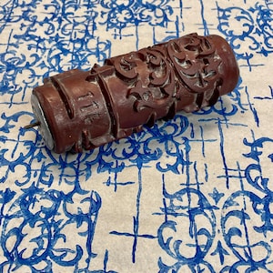 Può includere: Un rullo di legno intagliato, di colore marrone rossiccio scuro, con disegni floreali e a volute ornati. Il rullo ha un perno di metallo e una piccola ruota a un'estremità. Lo sfondo è una carta a motivi blu e bianca.