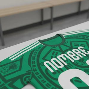 May include: Green soccer jersey with white accents and the word "Nombre" in large white letters. The jersey features a round neck and white stripes on the shoulders. The design includes Aztec-inspired patterns.